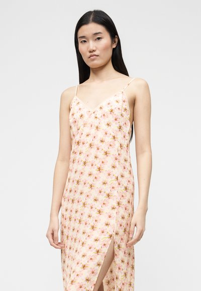 Νεαρή γυναίκα με μακριά σκούρα μαλλιά, που φοράει ροζ αμάνικο φόρεμα με λουλουδάτο μοτίβο, λεπτές τιράντες και σκίσιμο μέχρι τον μηρό.