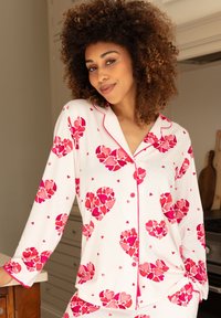 Witte pyjama set met een hartpatroon in roze en rood, met roze bies langs de kraag en de knopenlijst. Lange mouwen.
