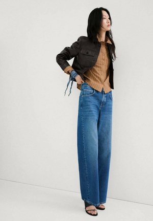 Mujer con camisa de rayas marrones, chaqueta negra corta, vaqueros de pata ancha azules y tacones abiertos negros, de pie contra un fondo claro y liso.