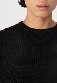 Suéter negro de punto con textura de canalé y cuello redondo, mostrando una vista cercana de la parte superior del pecho.