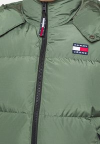 Zelená prošívaná bunda s vysokým límcem, zapínáním na zip a malým vyšívaným logem "Tommy Jeans" na hrudi. Texturovaná, lehká tkanina.