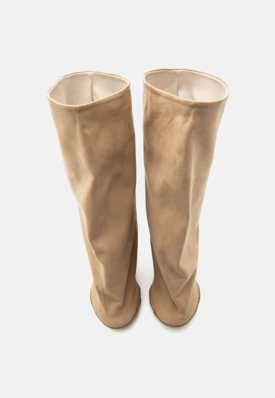 Ζεύγος ψηλών, μπεζ σουέτ μπότες με μαλακούς, χαλαρούς άξονες και στρογγυλεμένα δάχτυλα, τοποθετημένες όρθιες σε λευκό φόντο.