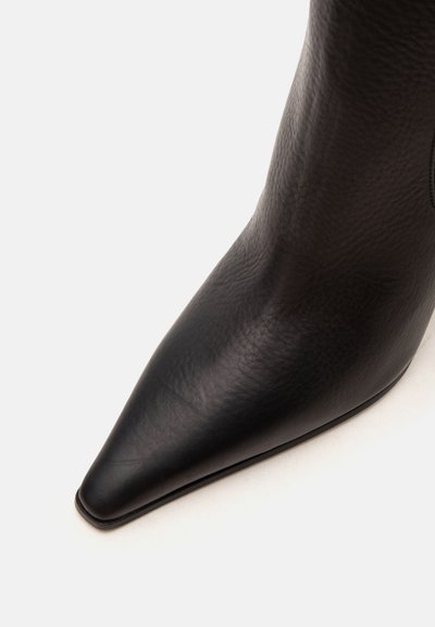 Gros plan d'une botte en cuir noir à bout pointu avec une surface lisse légèrement texturée sur un fond blanc.