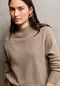 Beige Strickpullover mit geripptem Stehkragen und langen Ärmeln. Weiche Textur mit feinen Nähten entlang der Schultern.