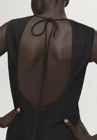 Vestido negro con un profundo corte en la espalda, sujeto con una delgada cinta. La textura suave de la tela contrasta con la piel. Presenta elementos de diseño minimalistas.