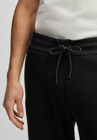 Svarta bomullsbyxor med elastiskt midjeband, med dragsko och sidofickor, uppvisade på en hand mot en neutral bakgrund.