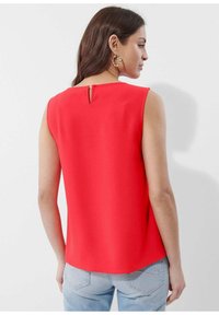 Rode mouwloze top met een keyhole-detail op de rug, een soepele textuur en een afgeronde zoom. Gecombineerd met lichtblauwe denim jeans op de achtergrond.