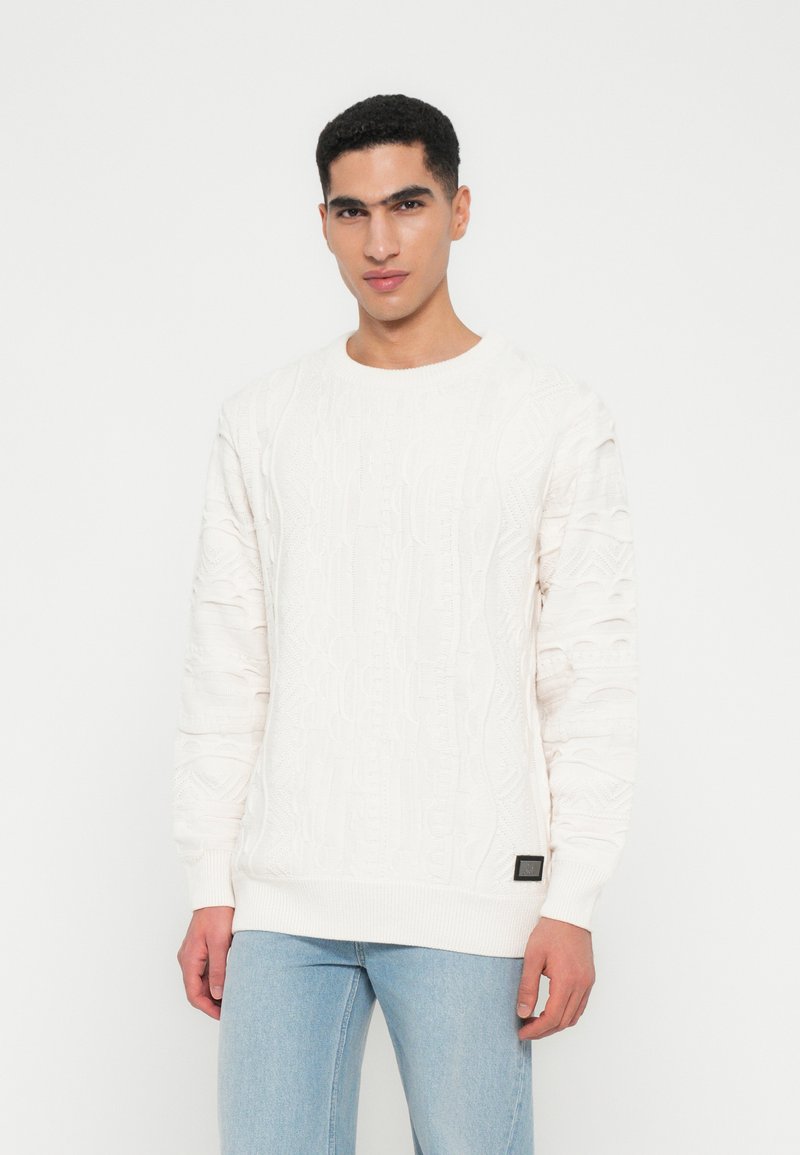 Jonge man in een wit gestructureerd gebreid trui en lichtblauwe jeans, staand tegen een effen witte achtergrond.