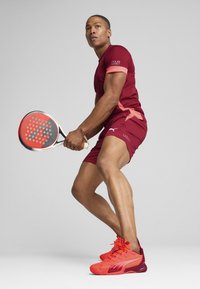 Rød atletisk skjorte med lyserøde accenter, røde shorts og klare røde sneakers. Spilleren holder en padelracket med et perforeret hoved.