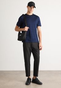 Jovem em uma t-shirt azul, calças pretas, ténis pretos e boné, segurando uma mala preta de ombro contra um fundo branco liso.