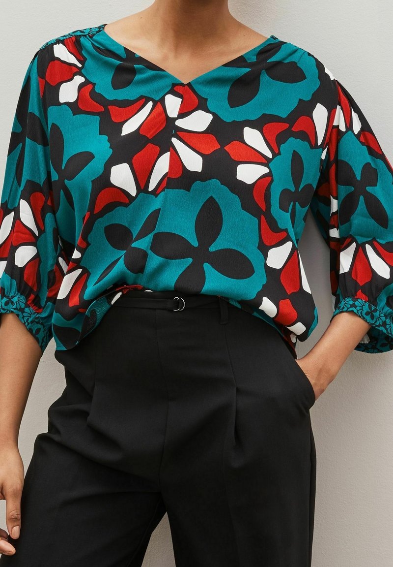 Blusa fluida color teal con motivo floreale nero, rosso e bianco, scollatura a V e maniche lunghe fino al ginocchio, abbinata a pantaloni neri a vita alta.