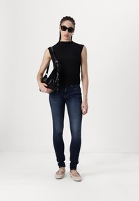 Melnā bez piedurknēm tops, tumši zilie pieguļošie džinsi, krēmkrāsas plakanas kurpes ar siksnām un melna plecu soma ar sudraba furnitūru. Modelis stāv taisni.