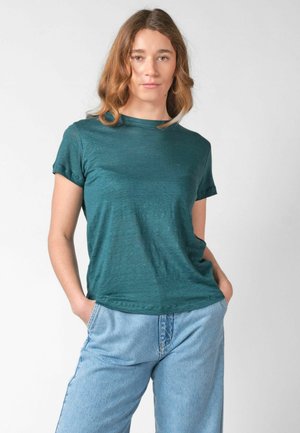 Fiatal nő hullámos barna hajjal, türkiz rövid ujjú pólót és világoskék magas derekú farmert visel, kezét a zsebében tartva áll.