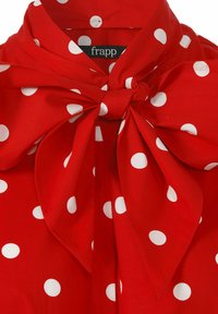 Blouse rouge avec de grands pois blancs, dotée d'un détail nœud au niveau du col. Tissu doux avec une texture lisse. Étiquette de marque visible.