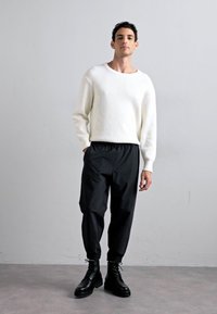 Valkoinen ribattuja neuletta, jossa on leveä kaula-aukko, yhdistettynä mustiin kapeneviin housuihin ja mustiin nauhakenkiin, yksinkertaista taustaa vasten.