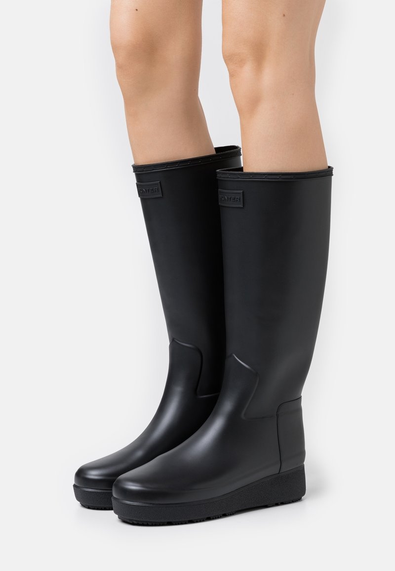 Bottes hautes en caoutchouc noir portées sur des jambes nues, posées sur une surface blanche unie avec une légère ombre en dessous.