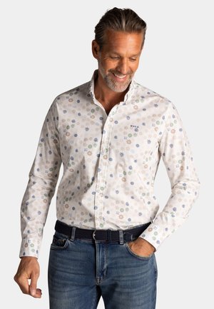 Man smiling, wearing a white button-up shirt with a small floral pattern and blue jeans, hand in pocket, against plain background.