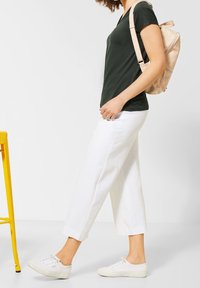 Top de manga corta verde oscuro, pantalones cortos blancos y zapatillas blancas. Mochila de color beige claro con detalle texturizado. Atuendo simple y casual.