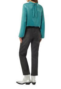 Türkise Satinbluse mit einem Bindebandverschluss, kombiniert mit dunkelgrauen Jeans im geraden Schnitt und silbernen Knöchelstiefeln. Durchgängige glatte Textur.