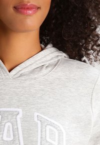 Grijze hoodie met een gestructureerde stof, verstelbare capuchon en witte geborduurde letters op de voorkant. Close-up van de halslijn.