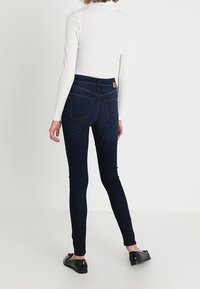 Jeans de color azul oscuro, elásticos y con una textura suave, de cintura alta y con bolsillos traseros. Llevados con una blusa ajustada de mangas largas blanca y bailarinas negras.