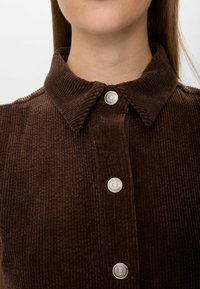 Rjava kordurojska srajca s ovratnikom in srebrnimi pritiski, ki ima rebrasto teksturo in bližnji pogled na ovratnik.