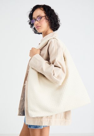 Grande borsa tote a quadri bianco sporco con un motivo testurizzato, indossata sopra un giubbotto beige chiaro, che completa un paio di pantaloni di jeans corti casual.