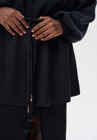 Czarny strój z teksturowanego, miękkiego materiału. Posiada pleciony ściągacz z złotymi akcentami oraz długie rękawy, prezentujące nowoczesny design.