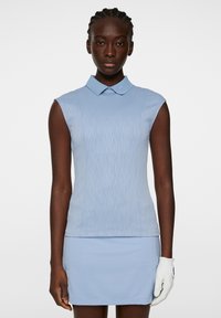 Vaaleansininen ilman hihoja oleva golfpaita, jossa on tekstuuripintainen kuvio, litteä kaulus ja napakka muotoilu, yhdistettynä sävyyn sopivaan hameeseen. Valkoinen golfkäsine käytössä.