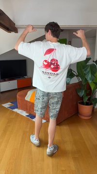 Persoon in camo shorts en grijze sneakers wijst met duimen naar een wit Adidas-shirt met rood kersen ontwerp, staand in de woonkamer met plant en tv.