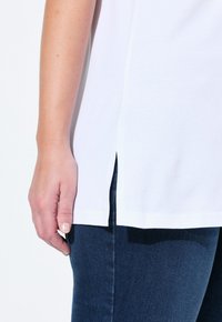 T-shirt en coton blanc avec fentes sur les côtés, associé à un denim bleu foncé. Gros plan sur la manche et la main, mettant en avant la texture du tissu et les détails de coupe.