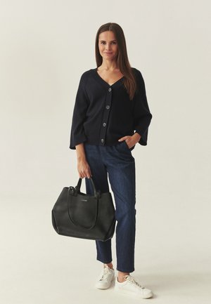 Žena v černém kardiganu, tmavých džínách a bílých teniskách držící velkou černou kabelku, stojící před jednobarevným pozadím.