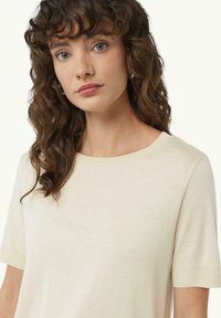 Kremowa koszulka z krótkim rękawem, wykonana z gładkiego materiału, z okrągłym dekoltem i subtelną fakturą, noszona przez modelkę z falującymi włosami.