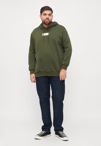 Sudadera de color verde oliva con un logo en el frente, cordones, puños de canalé, combinada con jeans oscuros y zapatillas negras con suelas blancas.