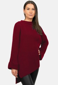 Pull en tricot bordeaux avec une texture côtelée, des manches larges et un ourlet asymétrique. Associé à des leggings noirs, il met en valeur une coupe décontractée.