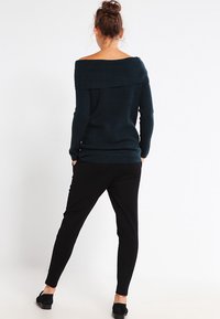 Ciemnozielony sweter typu off-the-shoulder z prążkowanym wzorem. Połączony z czarnymi, zwężającymi się spodniami. Całość dopełniają codzienne buty.