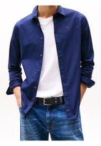 Camisa de botones color marino, tela de algodón, mangas largas enrolladas en los puños, usada sobre una camiseta blanca, combinada con jeans de mezclilla azul y un cinturón negro.