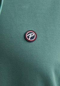 Grünes Baumwoll-T-Shirt mit einem runden schwarz-roten Logo-Patch auf der Brust, das ein stilisiertes 'P' zeigt. Glatte Textur, lässiges Design.