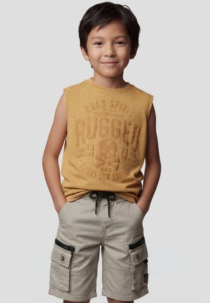 Niño joven de pie con las manos en los bolsillos, vistiendo una camiseta sin mangas mostaza con un gráfico de calavera y pantalones cortos cargo beige con cremalleras.