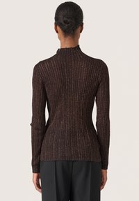 Kvinna med håret i en knut, klädd i en åtsittande, långärmad, ribbad mörkbrun topp med subtil lyster, står vänd bort från kameran framför en neutral bakgrund.