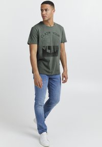 Olivenfarget bomull T-skjorte med teksten "CLAIM YOUR DENIM" i svart, kombinert med lyseblå jeans og hvite joggesko. Enkel design med rund hals.