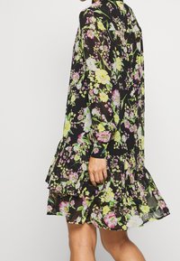 Sort blomstret kjole med levende pink og gule blomster, gennemsigtigt stof, lange ærmer og en lagdelt flæsekant.