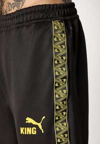Sorte sportsbukser med gult Puma-logo og tekst "KING" på låret, der har et mønstret gult geometrisk stribe langs siden.