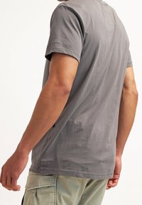 Šedé tričko s krátkým rukávem a volnějším střihem, s jemným logem na levém rameni a malou kapsou na horní části zad.