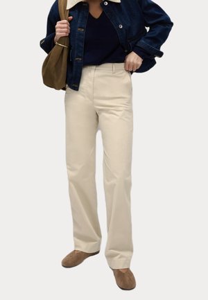 Person trägt beige weit geschnittene Hosen, navyfarbenes Oberteil, dunkle Jeansjacke mit weißem Kragen, braune Schuhe und trägt eine braune Umhängetasche.
