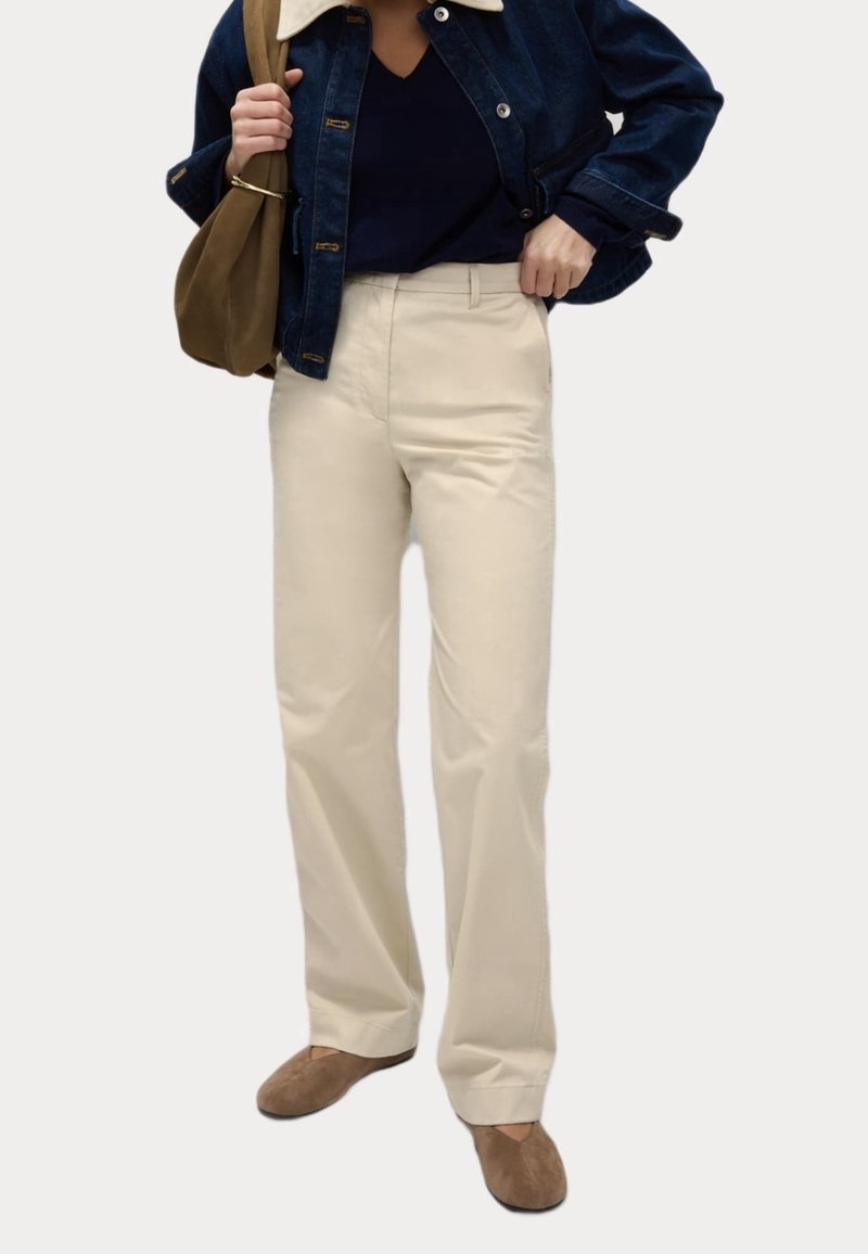 Personne portant un pantalon beige à jambes larges, un haut bleu marine, une veste en jean foncée avec un col blanc, des chaussures marron, et portant un sac à épaule marron.
