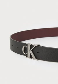 Zwarte leren riem met een gladde textuur, voorzien van een zilverkleurige CK-logo gesp. De binnenkant is bordeauxrood, met subtiele stikseldetails.