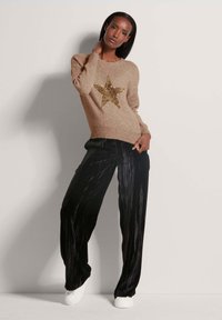 Beige gestrickter Pullover mit einem großen goldenen Paillettenstern-Design, kombiniert mit schwarzen plissierten Samthose und weißen Sneakers.