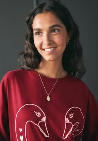 Burgundia sweatshirt valgete tikitud luikedega, millel on ümmargune kuldne ripatsiehe. Sile kangatekstuur, mugav lõige.