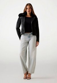 Veste en cuir noir avec un col doux, associée à un jean bleu clair à jambes larges et des sandales noires. Le design est ajusté et moderne.